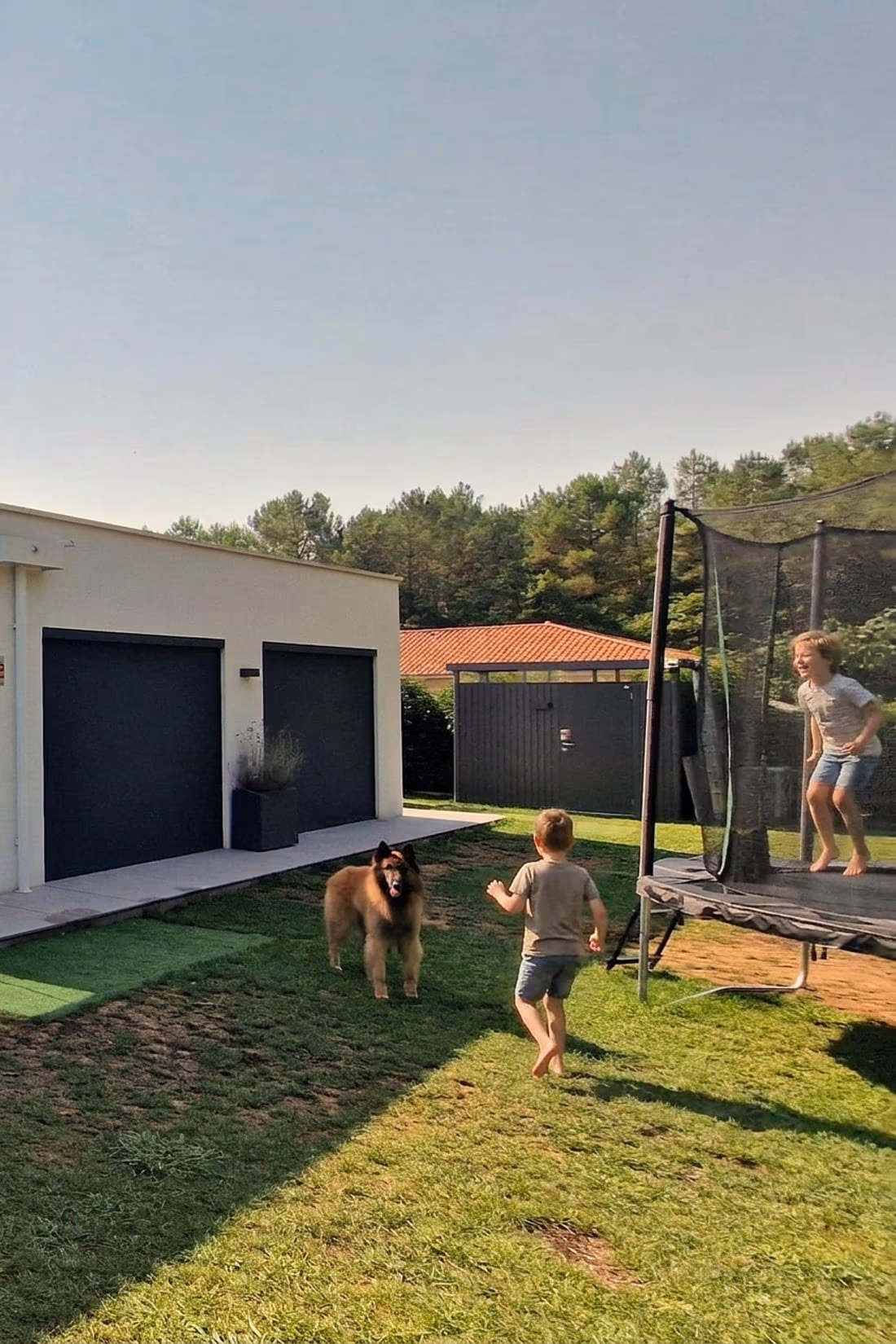 Enfants pieds nus qui jouent dans le jardin, sur la pelouse et au trampoline, devant une maison de plain-pied Landes Habitat à Saint-Vincent-de-Paul (40)