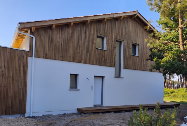 Façade de maison à étage avec bardage bois vertical, enduit blanc, fenêtres et terrasse en bois.