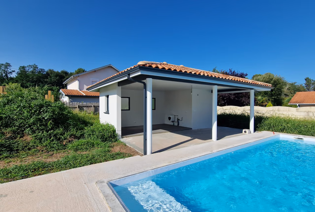 Pool-house blanc avec toiture en tuiles et piliers gris devant une piscine rectangulaire à l'eau bleue.