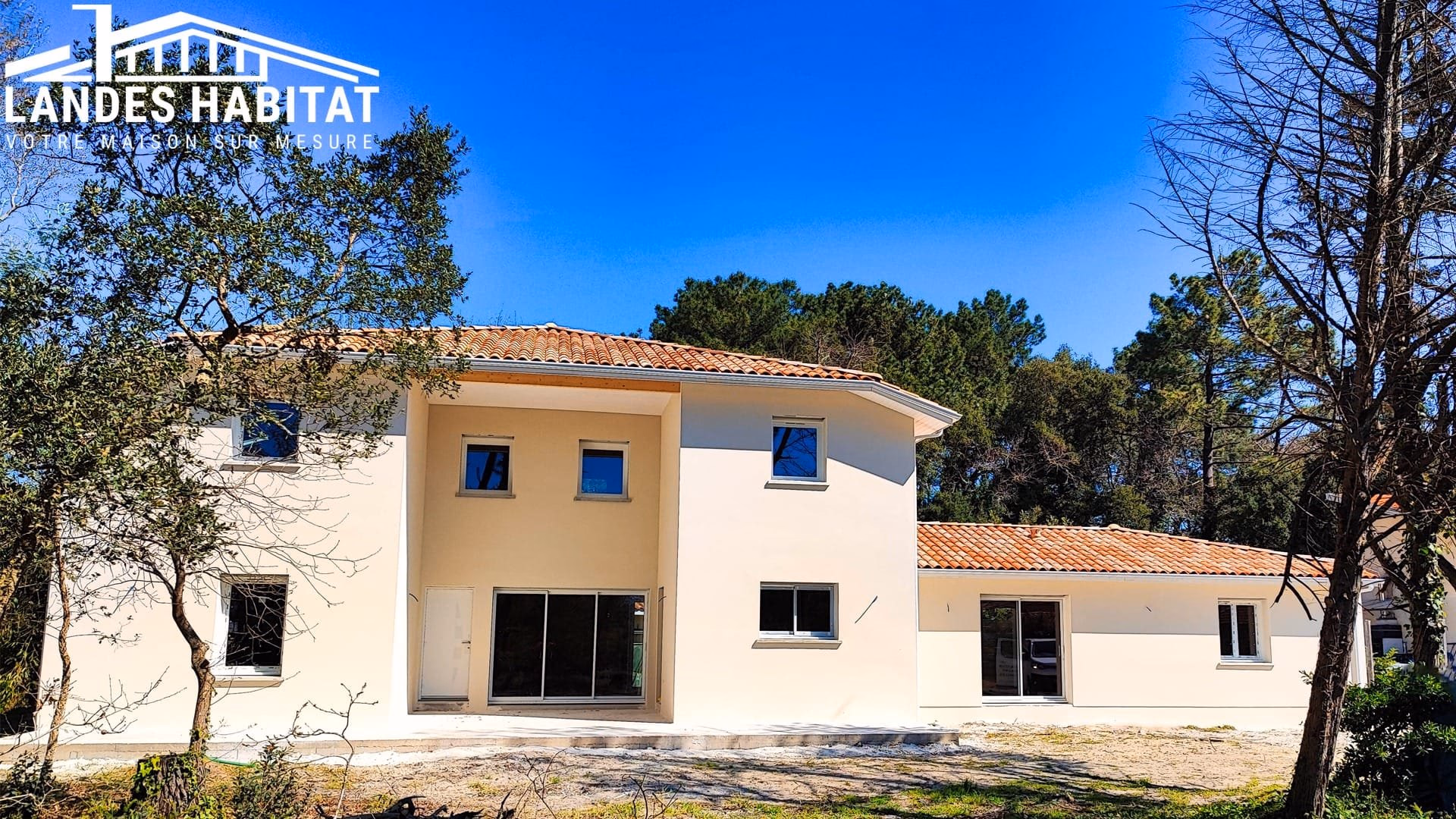 Maison individuelle neuve à étage avec enduit beige clair fini, toiture en tuiles et grand ciel bleu.