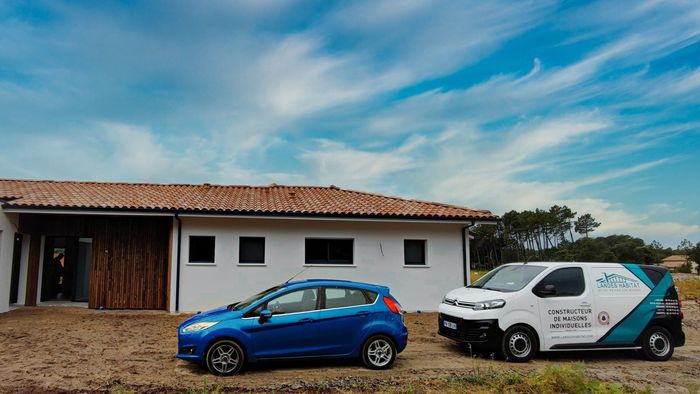 Voiture bleue et utilitaire blanc de constructeur stationnés devant une maison neuve aux tuiles rouges.