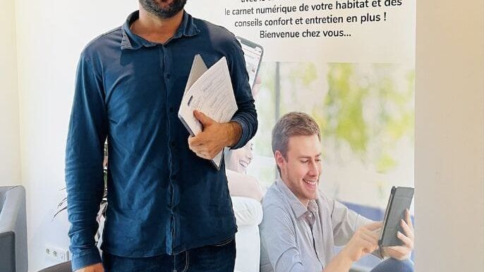 Un homme souriant debout tient des dossiers devant un roll-up promotionnel My Landes Habitat.