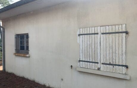 Mur extérieur d'une maison avec une fenêtre grillagée et des volets en bois fermés.